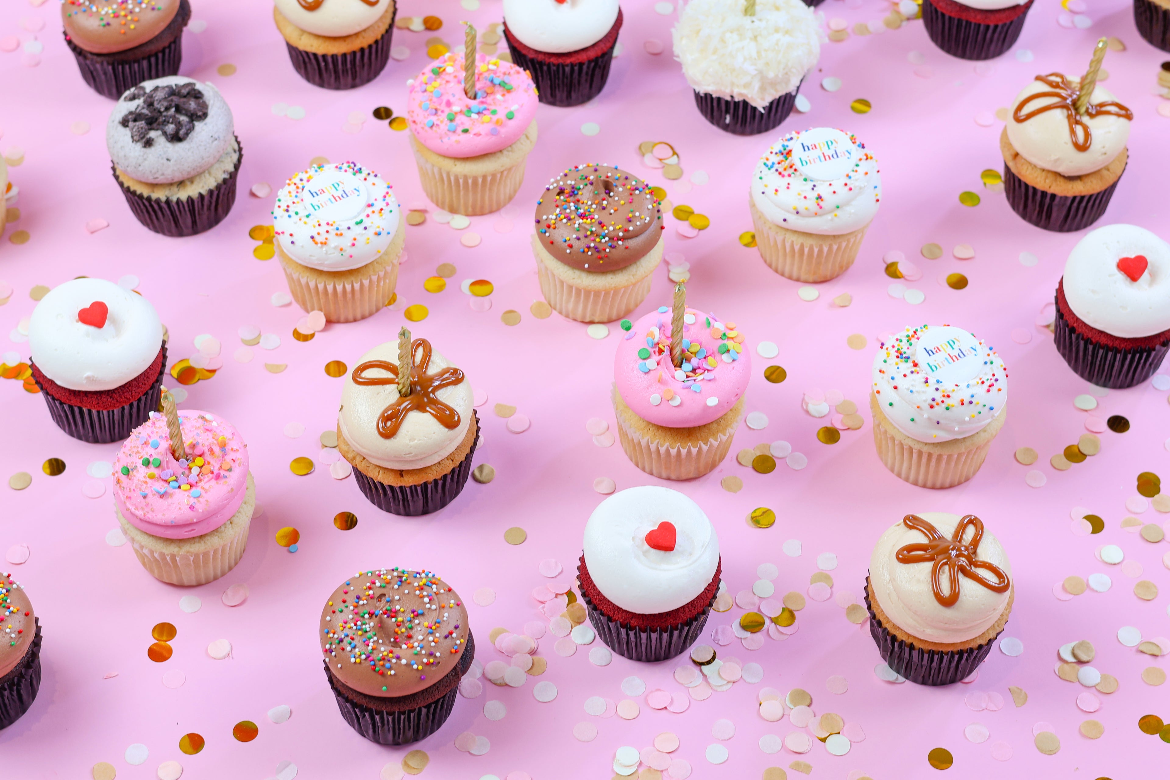 Festive assortment of decorated cupcakes with candles and confetti on a pink background